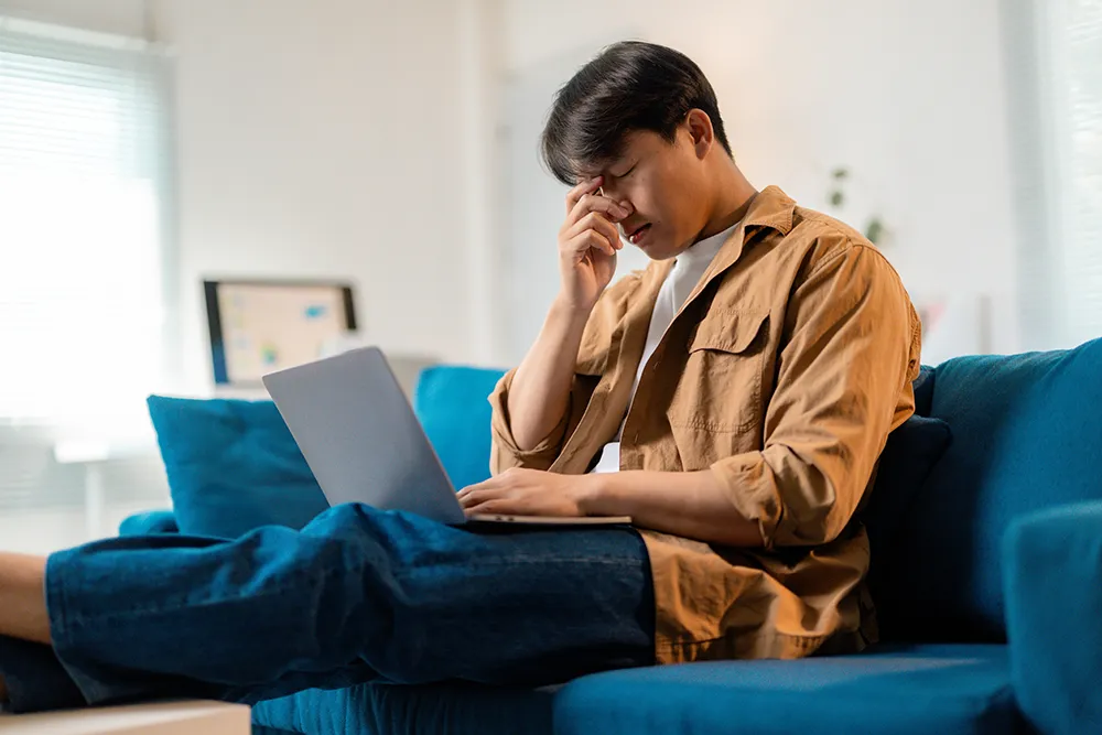 a man sitting on his sofa with his laptop and rubbing his eyes