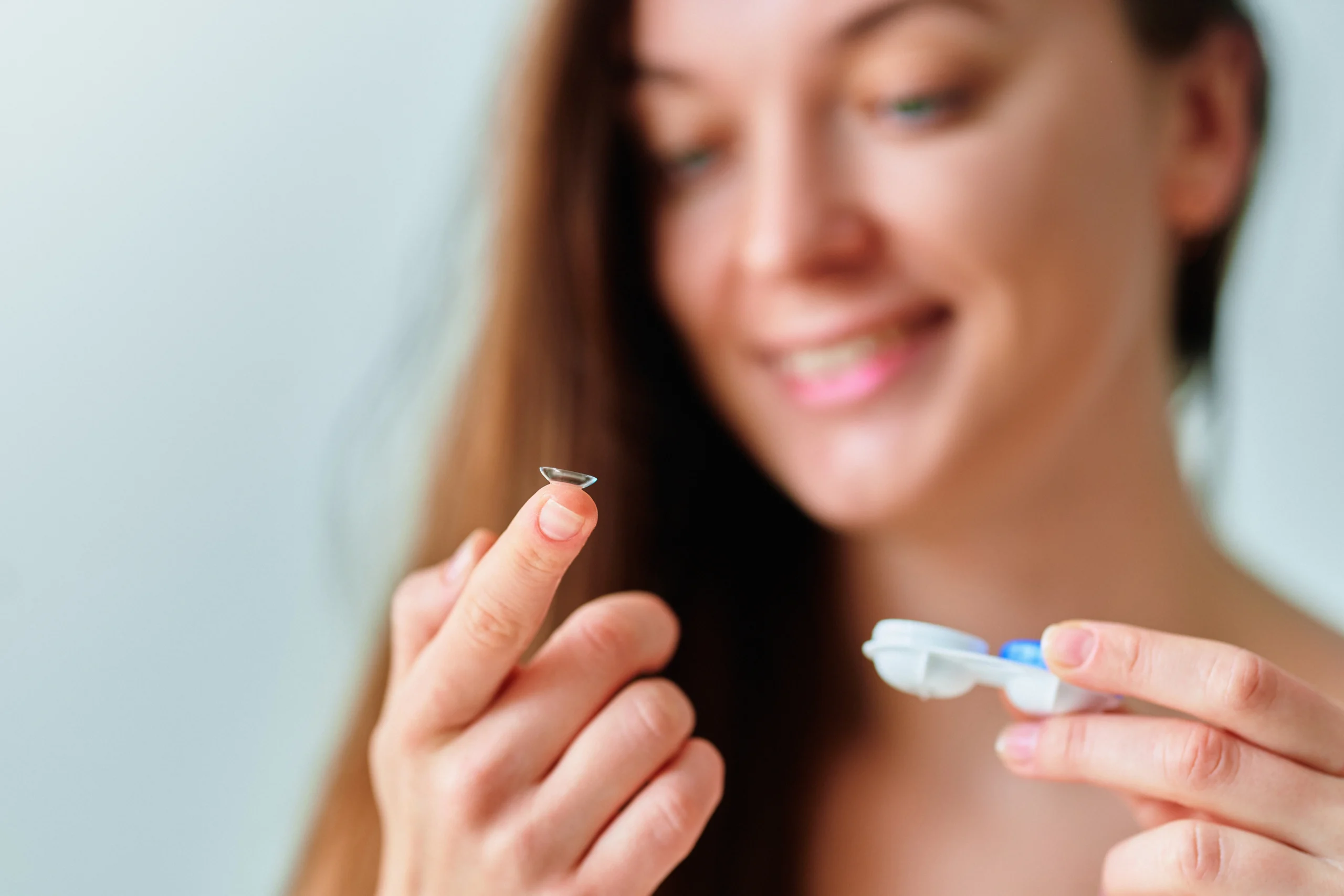 A smiling woman holding a contact lens case in one hand and resting a lens on her index finger.